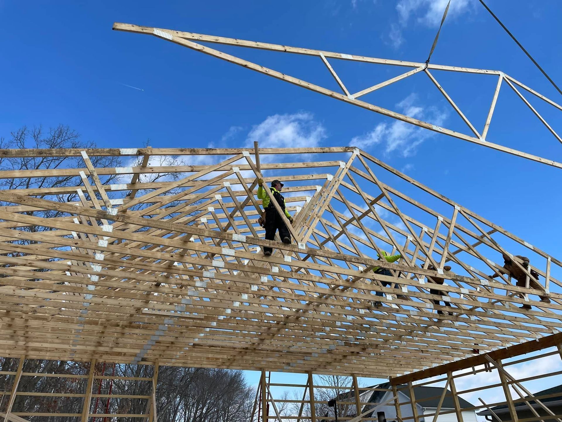 Workers building a wooden roof frame under a blue sky, with a crane lifting roof trusses above.