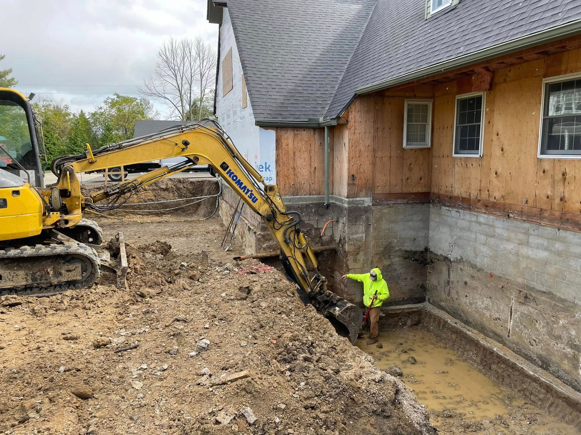 Yellow excavator digs a trench beside a house while a worker in a neon jacket stands in muddy water.