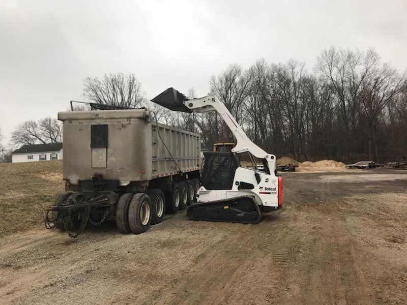 White skid-steer loader dumping material into a large gray dump truck at a muddy worksite