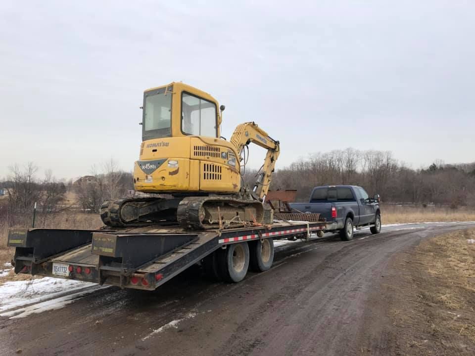 Yellow excavator on a flatbed trailer pulled by a black truck on a muddy rural road.