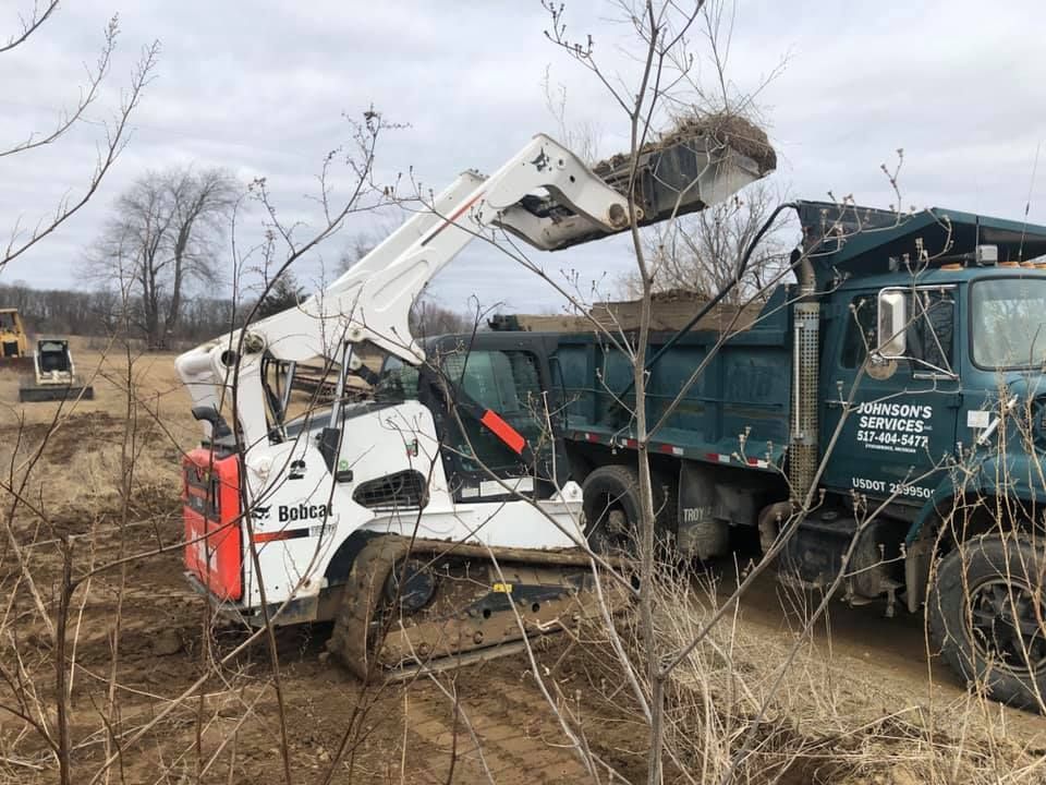 White skid-steer loader dumping debris into a green dump truck in a muddy field