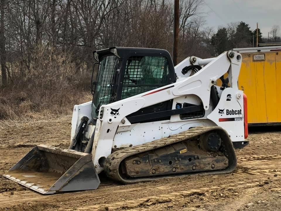 White Bobcat skid-steer loader on muddy ground with front bucket lowered in a wooded area