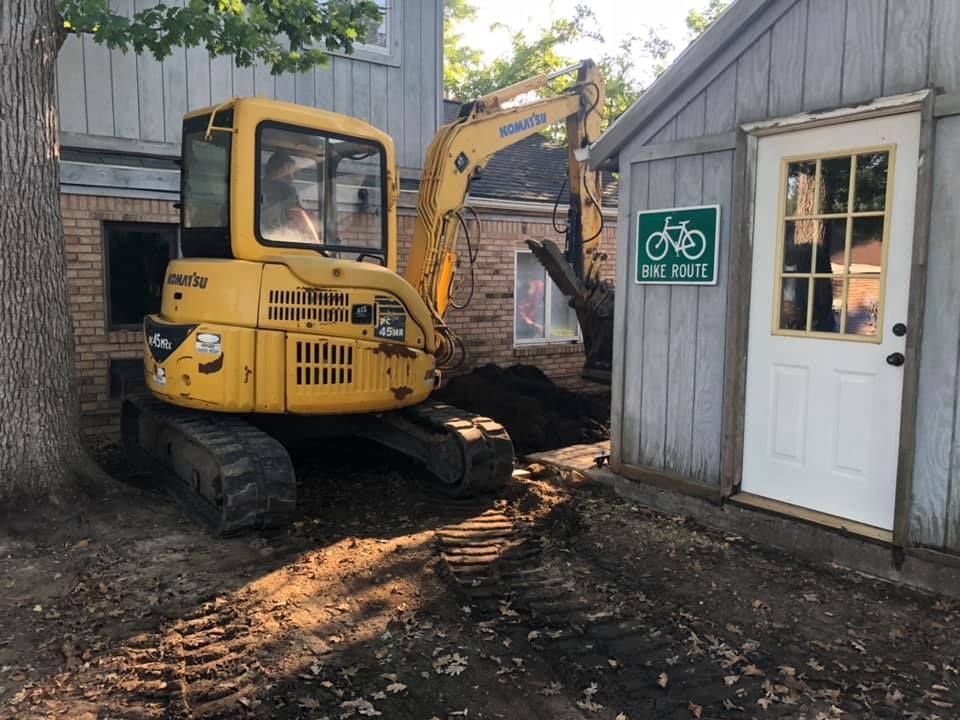 Yellow excavator parked beside a shed and white door with a green bike sign in a dirt yard.