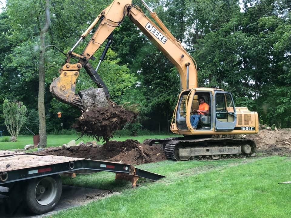 Yellow excavator lifting a root ball beside a flatbed truck on a grassy construction site