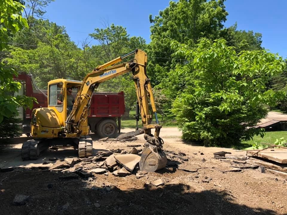 Yellow excavator clearing rubble beside a red dump truck on a sunny wooded roadside