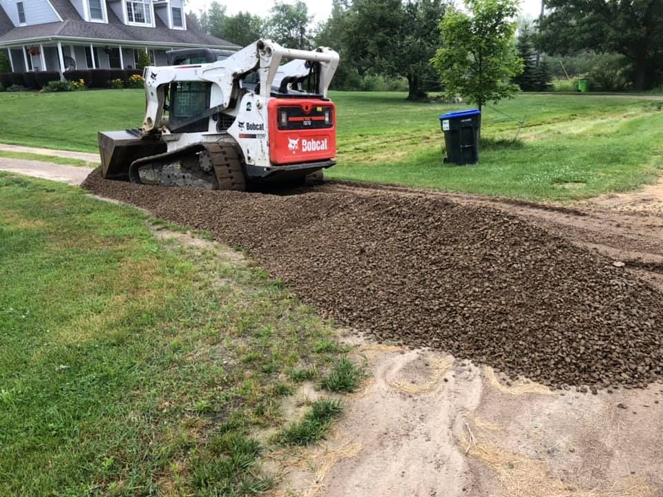 Skid steer spreading gravel on a dirt path beside a grassy yard and house