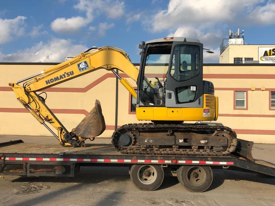 Yellow mini excavator on a flatbed trailer in an industrial yard under a cloudy sky