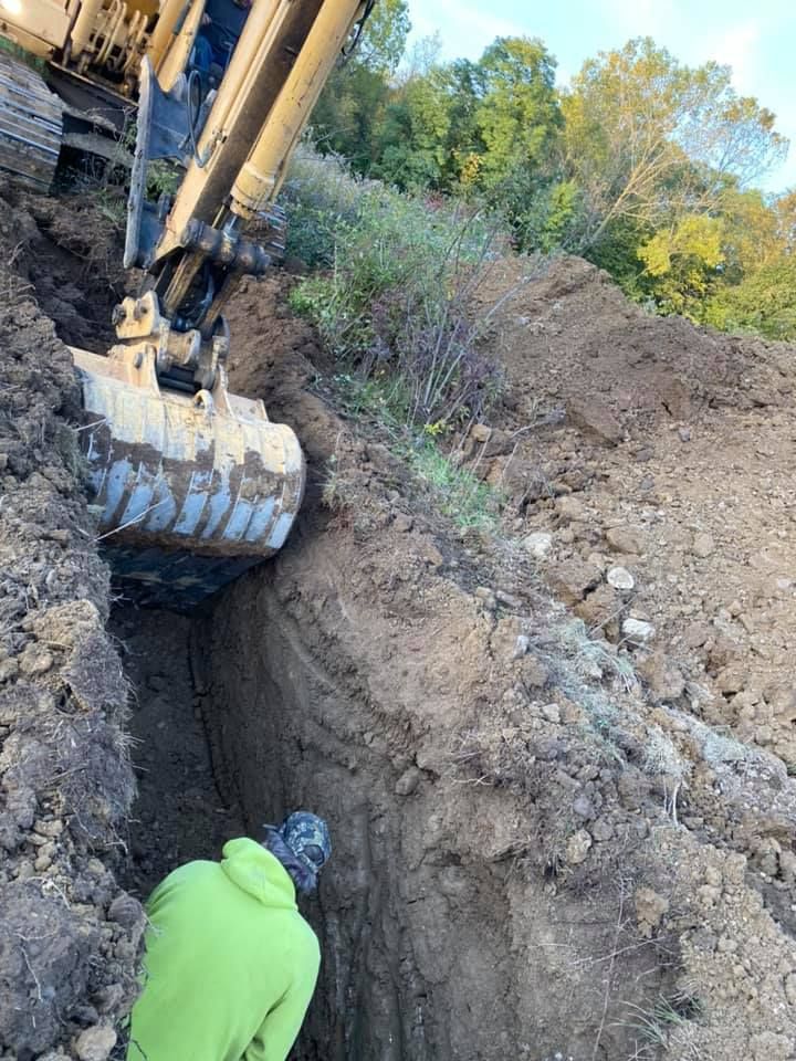 Construction excavator digging a deep trench beside a worker in a bright green hoodie.