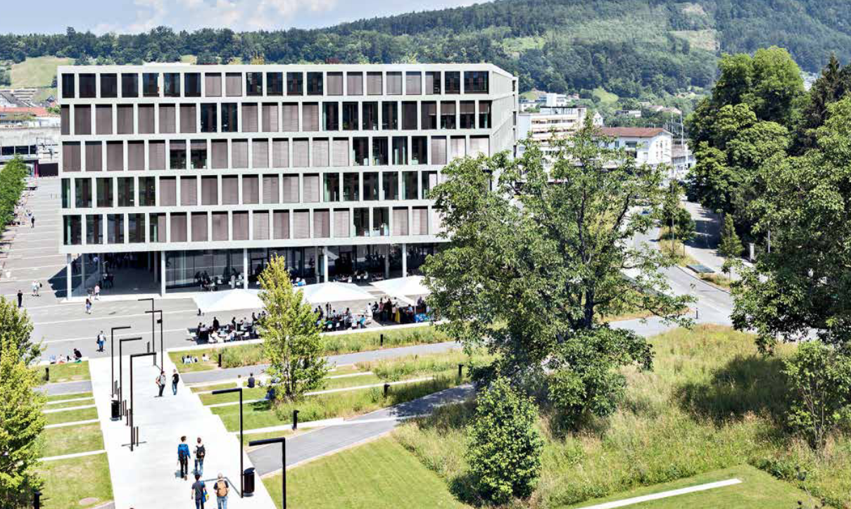 Ein grosses Gebäude mit vielen Fenstern ist von Bäumen und Gras umgeben.