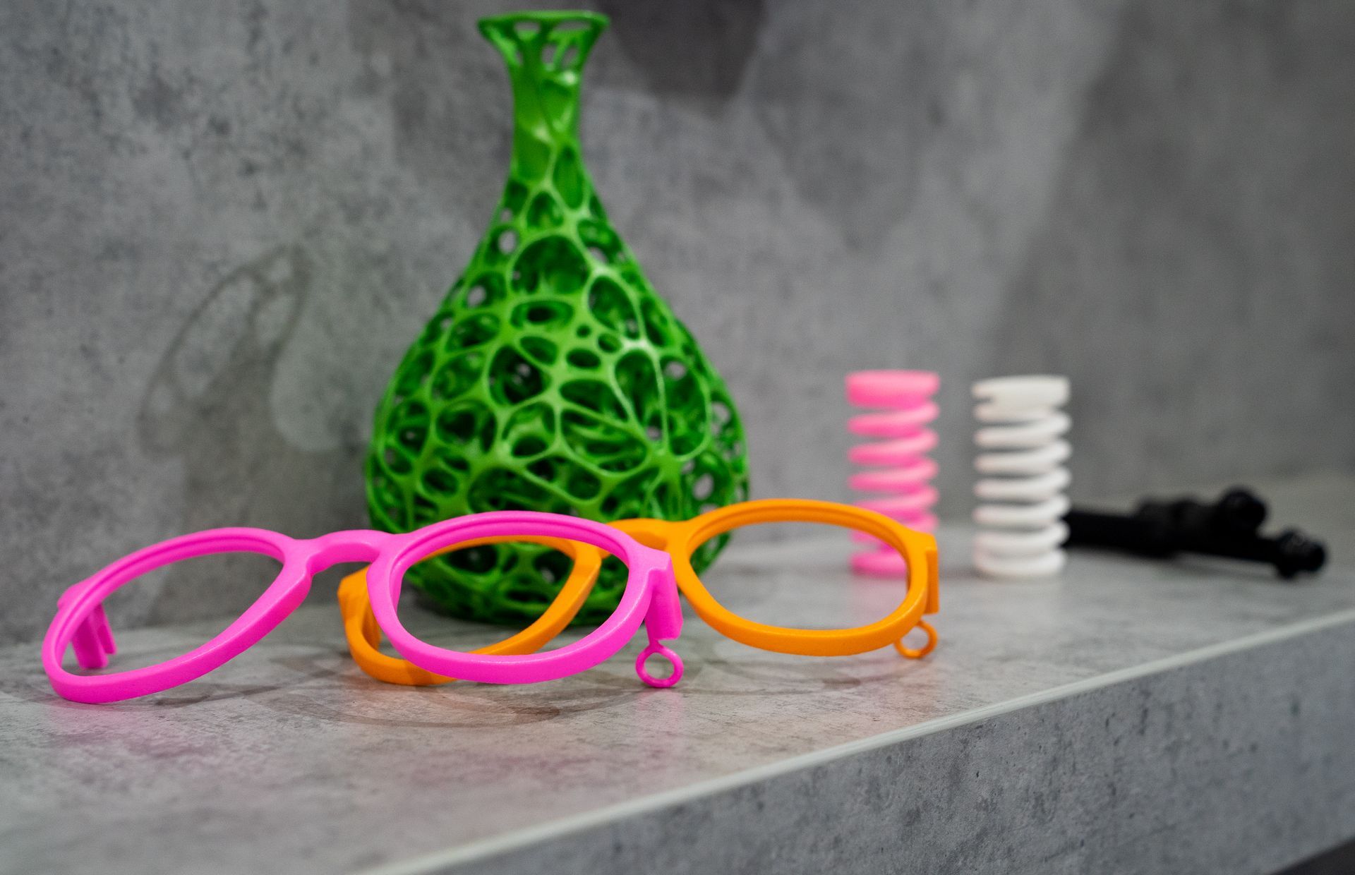 Eine rosa und eine orange Brille stehen auf einem Regal neben einer grünen Vase.