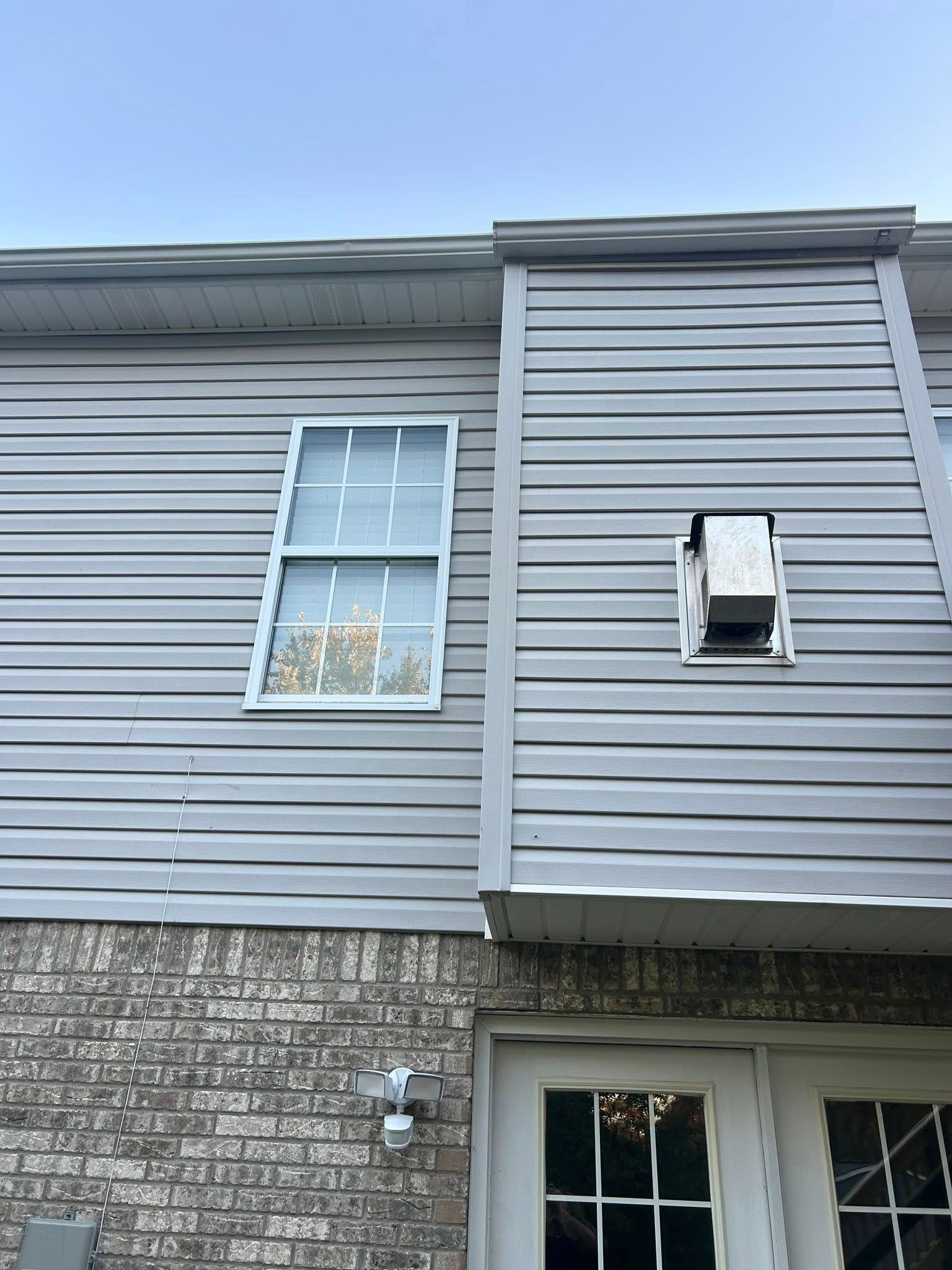 Exterior wall with grey siding, window, brick base, and a ventilation unit.