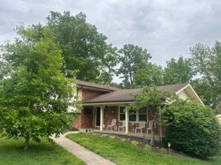 Suburban brick house with a front porch, surrounded by lush green trees and shrubs.