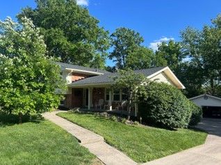 Single-story brick house with a front porch, green lawn, and trees under a blue sky Single-story brick house with a front porch, green lawn, and trees under a blue sky