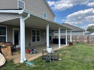 Covered backyard patio with white columns, tools, a chair, and a grassy yard beside a house.