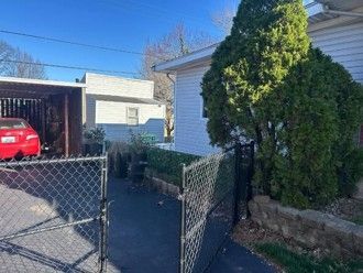Open chain-link gate leading to a driveway, with a red car in a garage and a white house beside a large evergreen tree. Open chain-link gate leading to a driveway, with a red car in a garage and a white house beside a large evergreen tree.
