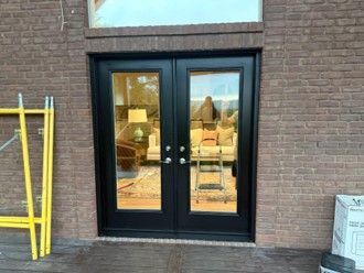 Black double doors in a brick wall, reflecting chairs and tables inside.