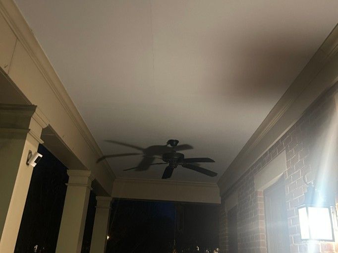 Covered porch at night with a ceiling fan and a lit wall lantern beside a dark doorway.