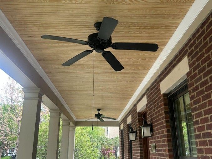 Covered brick porch with two black ceiling fans and a wood-paneled ceiling