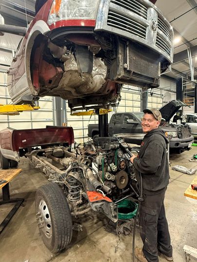 Mechanic beside a vehicle lifted above an exposed engine in a busy auto repair shop  | Drew's Diesel Repair