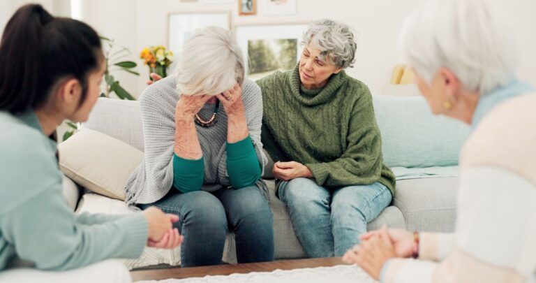 People sitting in a therapy group, one person crying while others listen supportively in a bright room