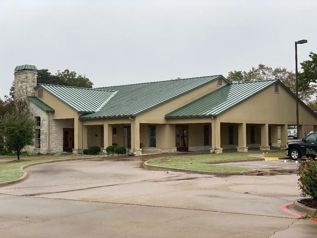 Single-story beige building with green metal roof and covered entrance beside a wet parking lot