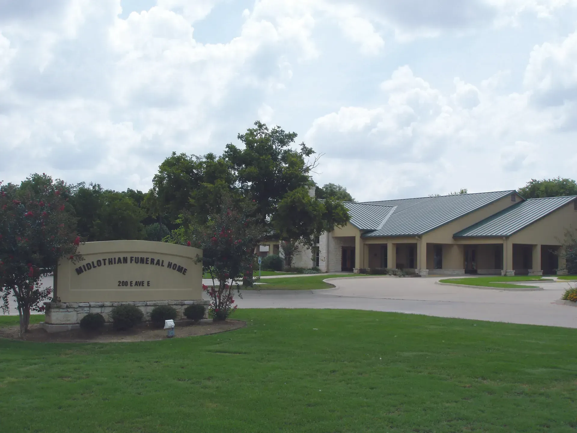 School building with a curved sign, green lawn, trees, and cloudy sky