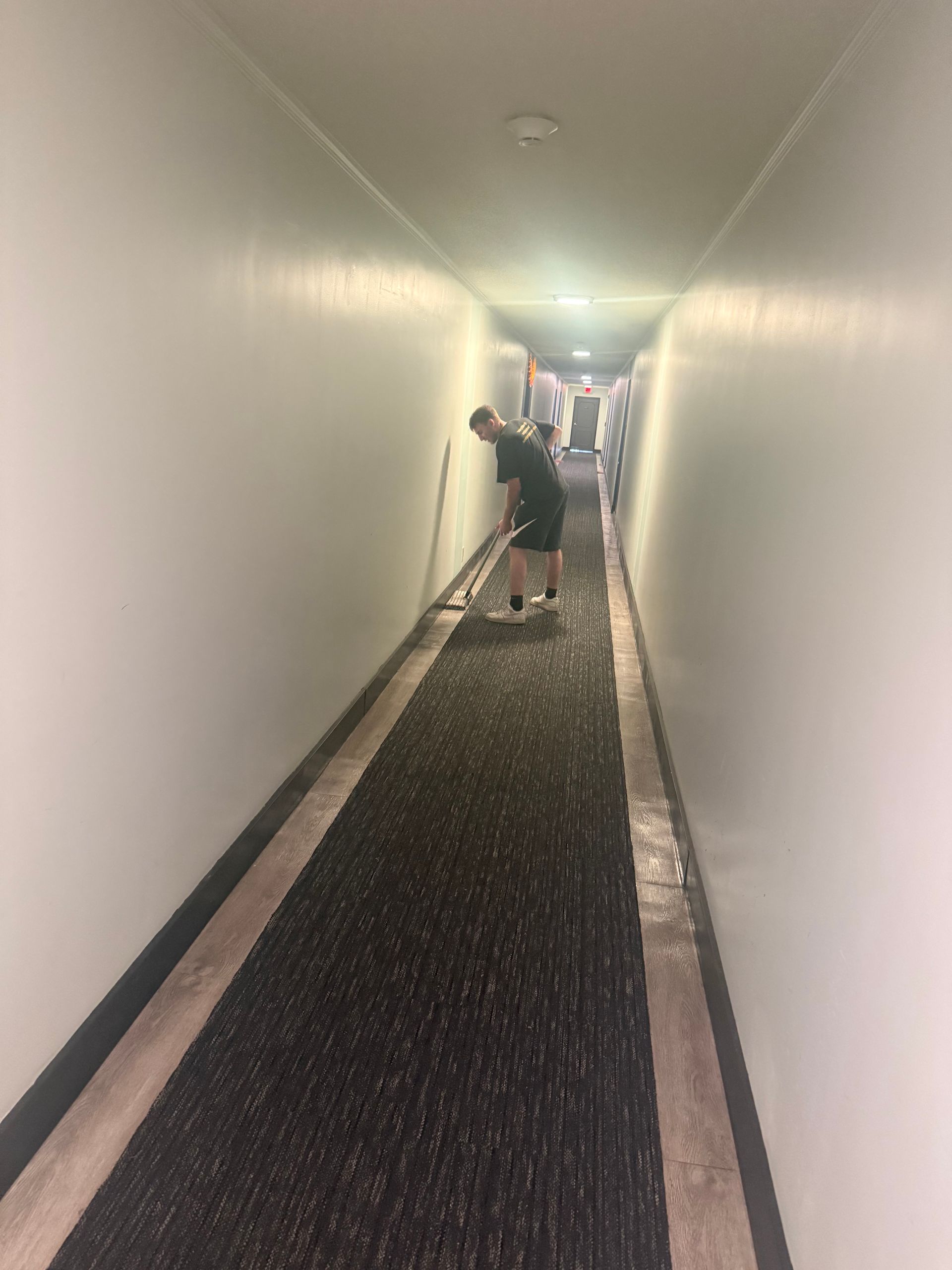 Person sweeping hallway with dark carpet and white walls.