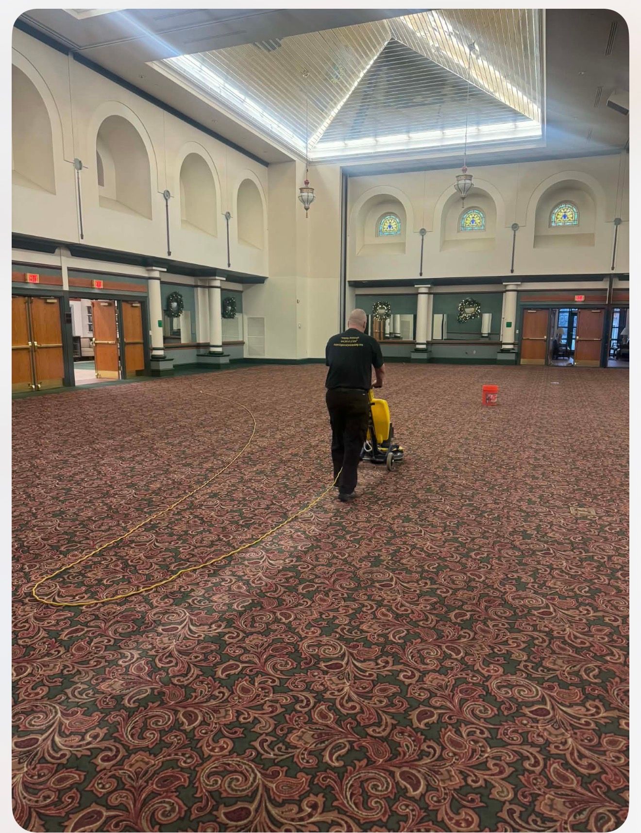 Person using a carpet cleaner in a large room with patterned carpet and arched windows.