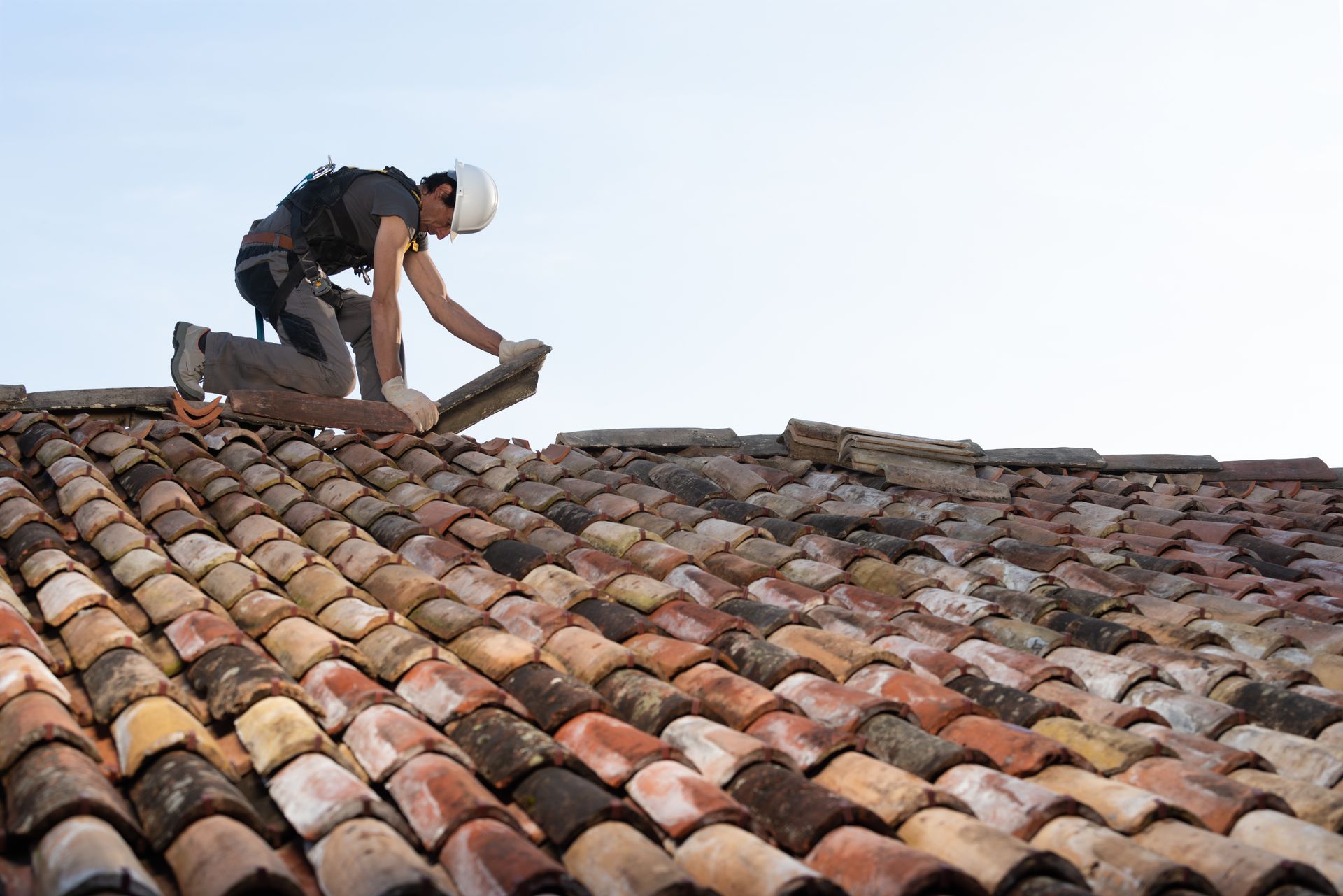 Residential roofing contractor in orange gear repairing tile roof at sunset with power tool. A roofer with a helmet and harness carefully installs clay tiles on a sloped roof.