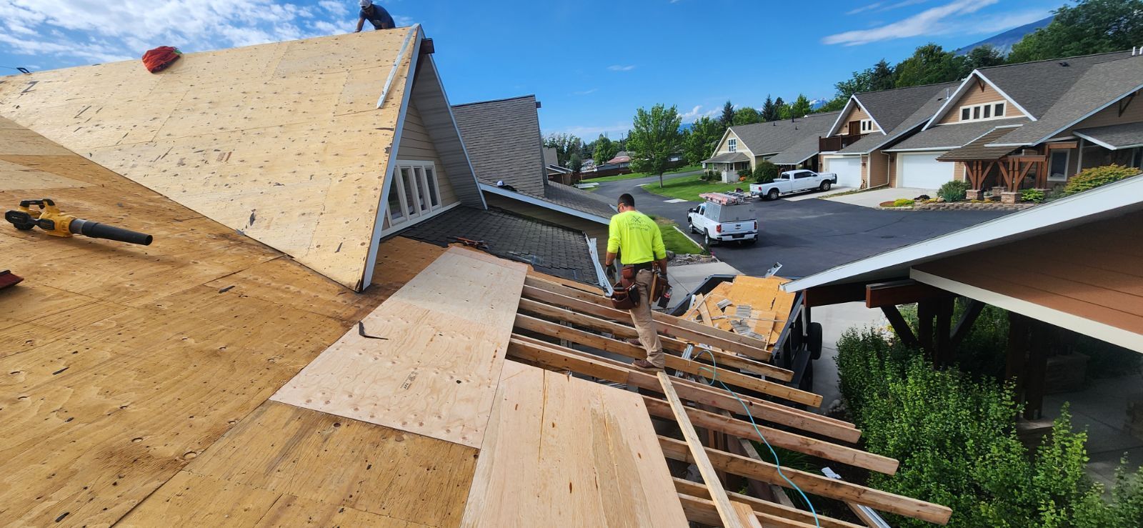 Roofers install plywood sheathing on a suburban home's roof under a bright, clear blue sky.