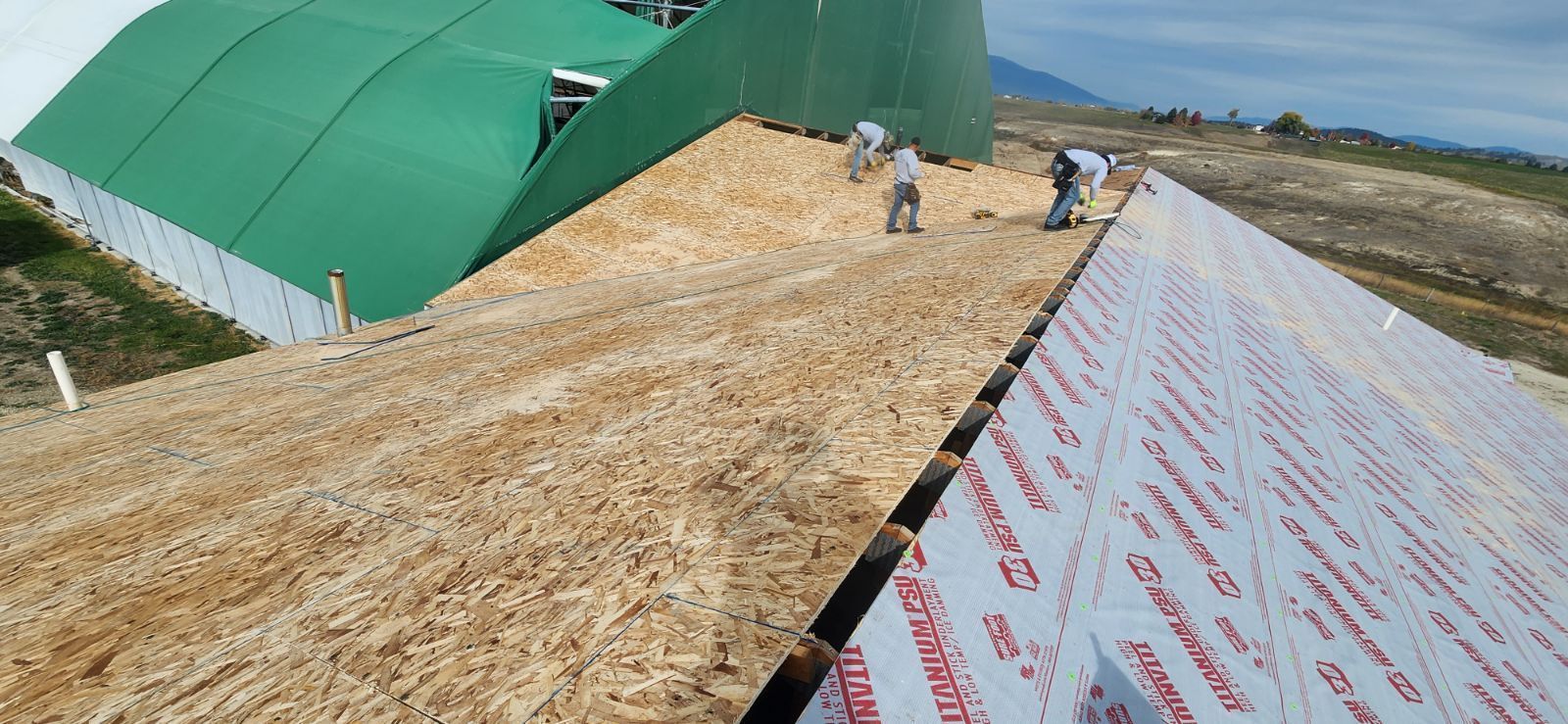 Construction workers installing white, branded roof sheeting on a building under a large, curved green cover.