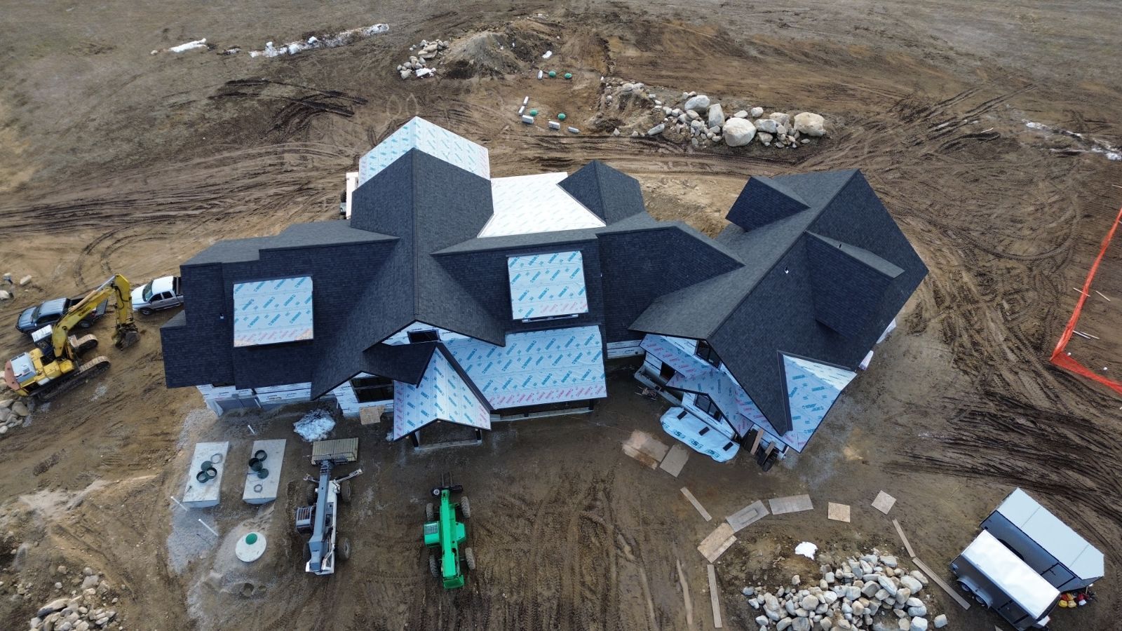 An aerial view of a construction site featuring a house with a complex, dark shingled roof and exposed blue roof sheathing.