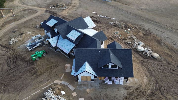 Aerial view of a new residential house under construction with a dark shingled roof, surrounded by bare dirt and equipment.