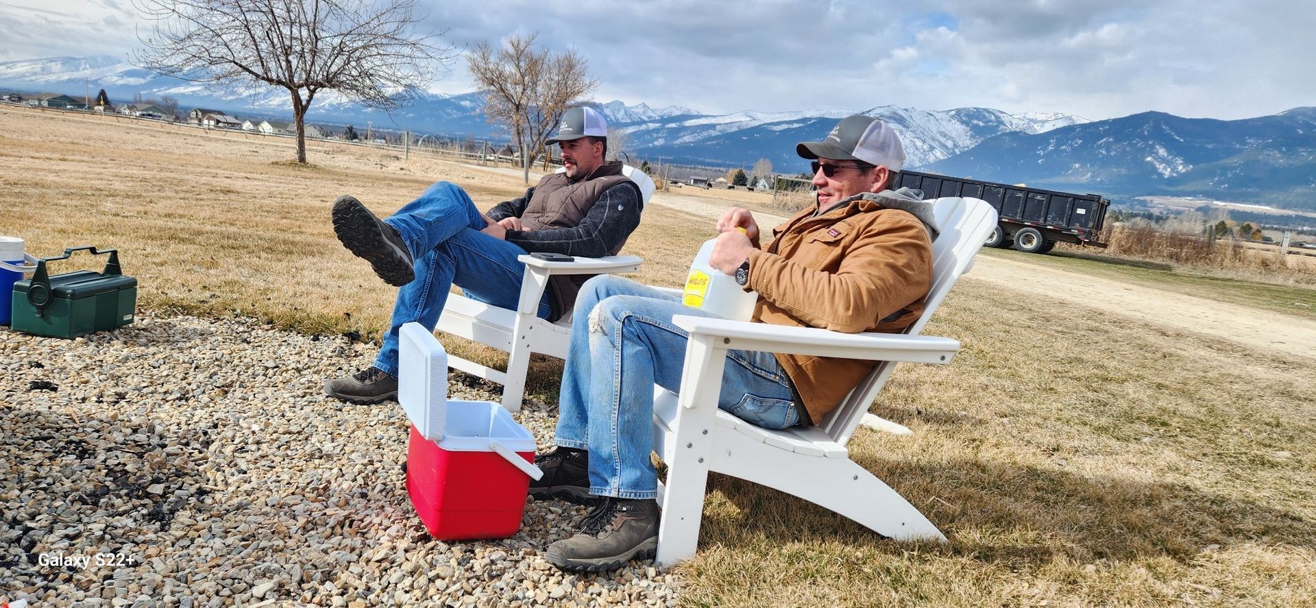 Two people wearing caps and jackets sit in white chairs outdoors, relaxing with a red cooler near a gravel area.