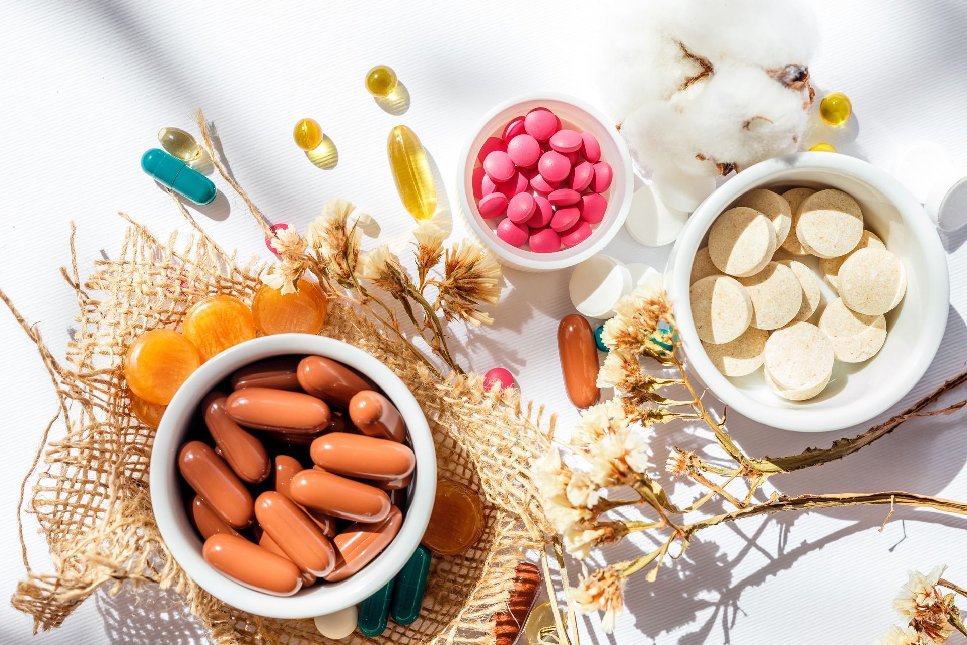Assorted capsules, tablets, and supplements in bowls on a bright white surface with dried flowers.