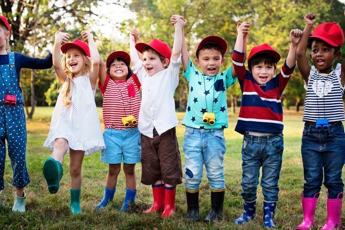 Children jumping with arms raised in a grassy park, wearing colorful hats and bright clothes