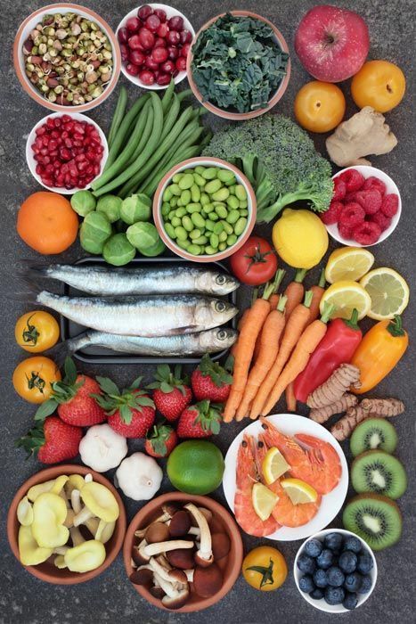 Assorted fresh fruits and vegetables arranged on a dark surface, including berries, greens, carrots, fish, and citrus.