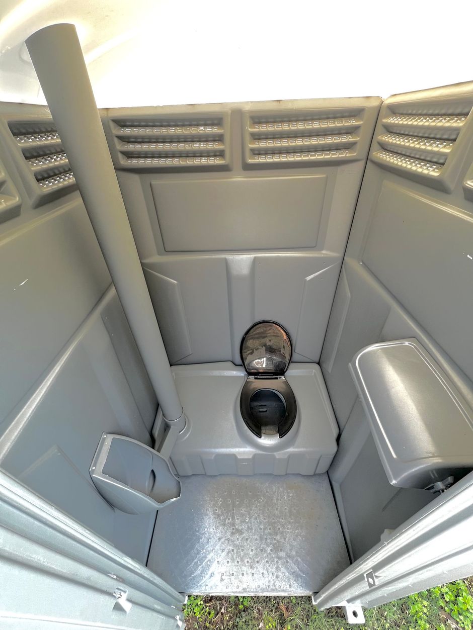 Interior of a portable toilet, grey plastic walls, open toilet seat, tall vertical vent pipe.