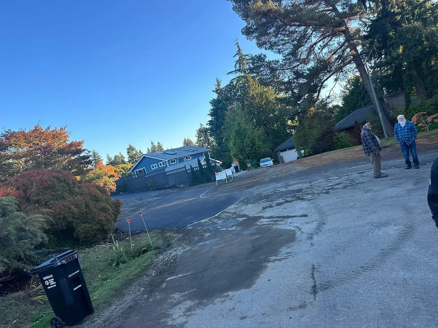 A sloped road with a large building, trees, and two people standing.  A trash can is in the foreground.