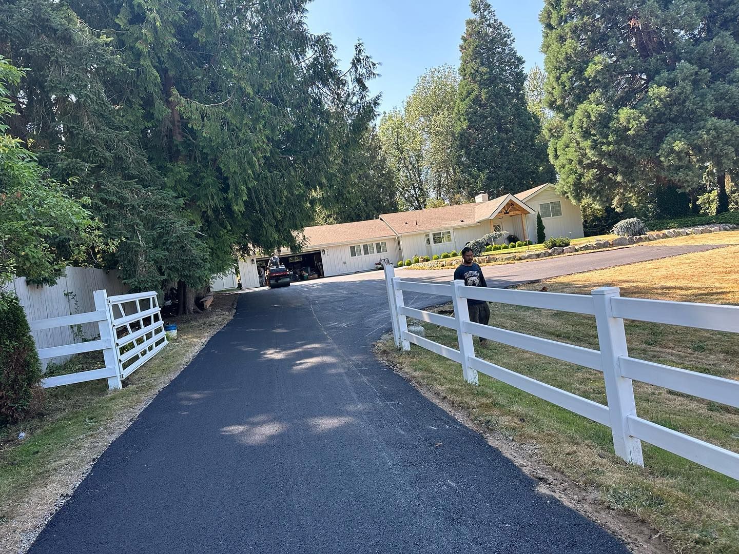 A long, black asphalt driveway leads to a white house with a partially visible fence on each side.