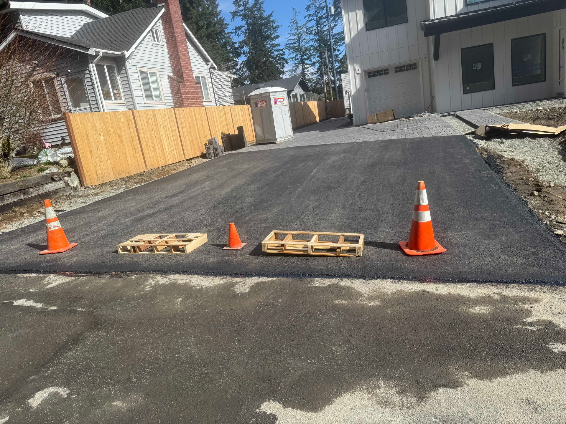 Asphalt paving in progress; worker operating roller near building with loading docks. Roadside cones present.