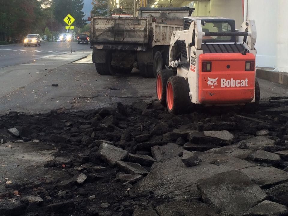 Bobcat skid-steer loader removing asphalt debris from a road. A dump truck waits nearby.