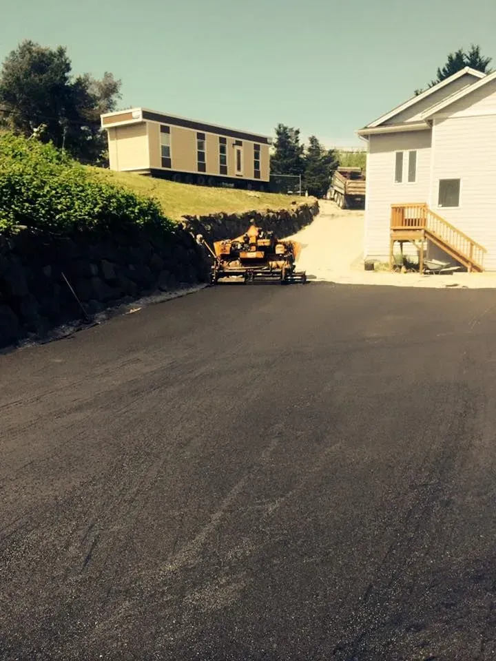 Asphalt driveway leading up a hill towards a modular home and a two-story building on a sunny day.