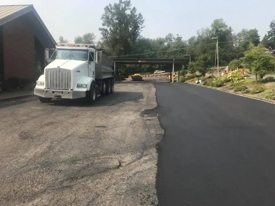 White dump truck next to a freshly paved driveway.  A building and trees are in the background.