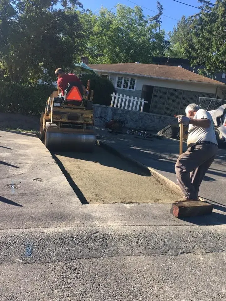 Construction workers paving a driveway: One operates a steamroller, the other compacts asphalt with a tool. Sunny day.