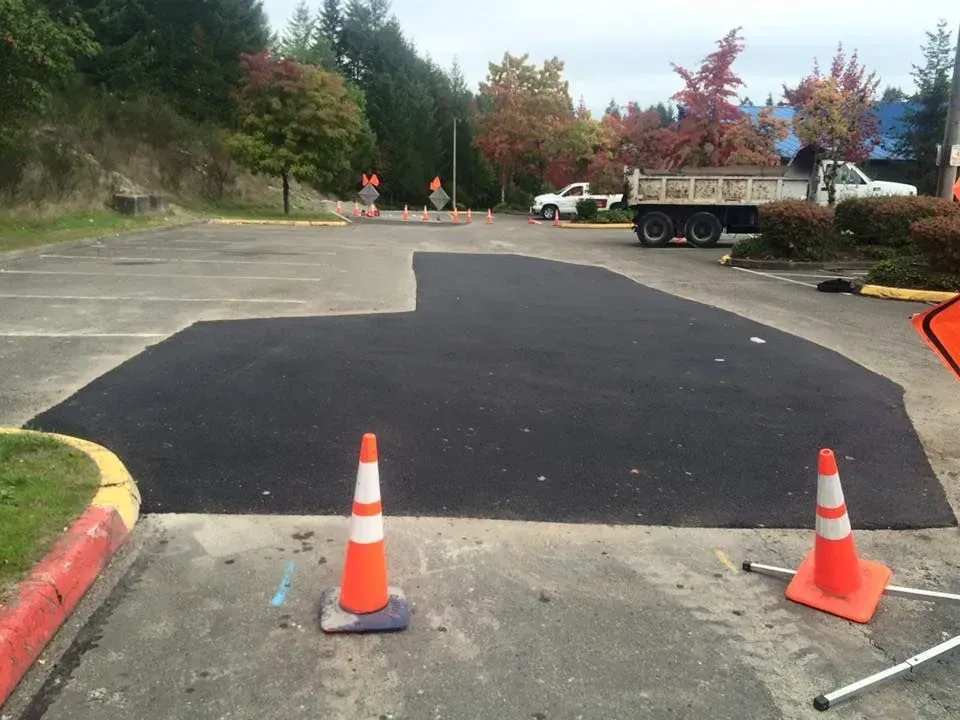 Paved area in a parking lot, with freshly laid asphalt surrounded by orange cones and a truck in the background.