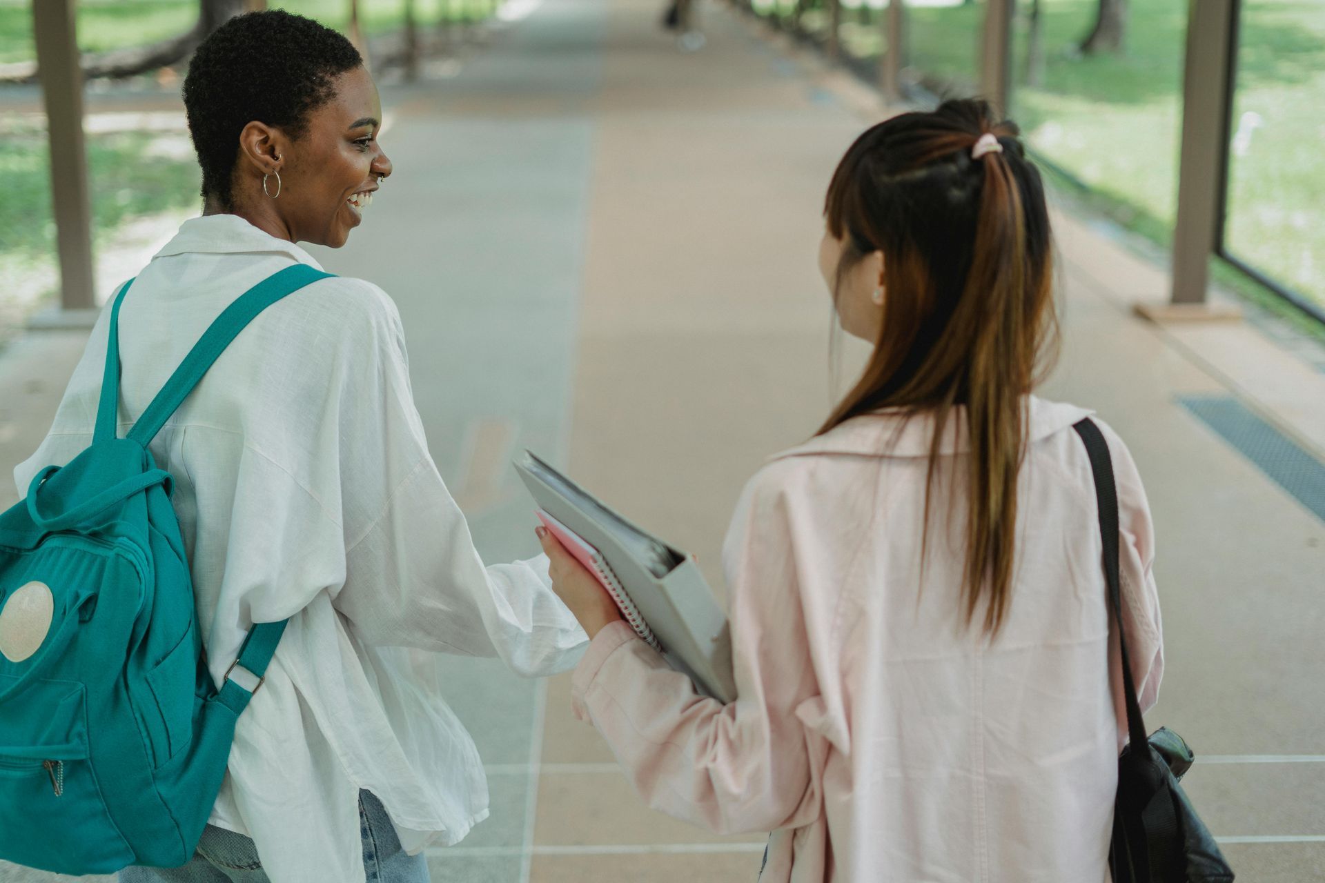 Two people walking and talking in a covered walkway, one with a backpack and one carrying a binder.