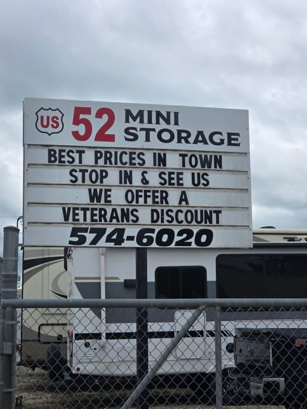 US 52 Mini Storage sign advertising best prices and veterans discount, with truck and fence below. US 52 Mini Storage sign advertising best prices and veterans discount, with truck and fence below.