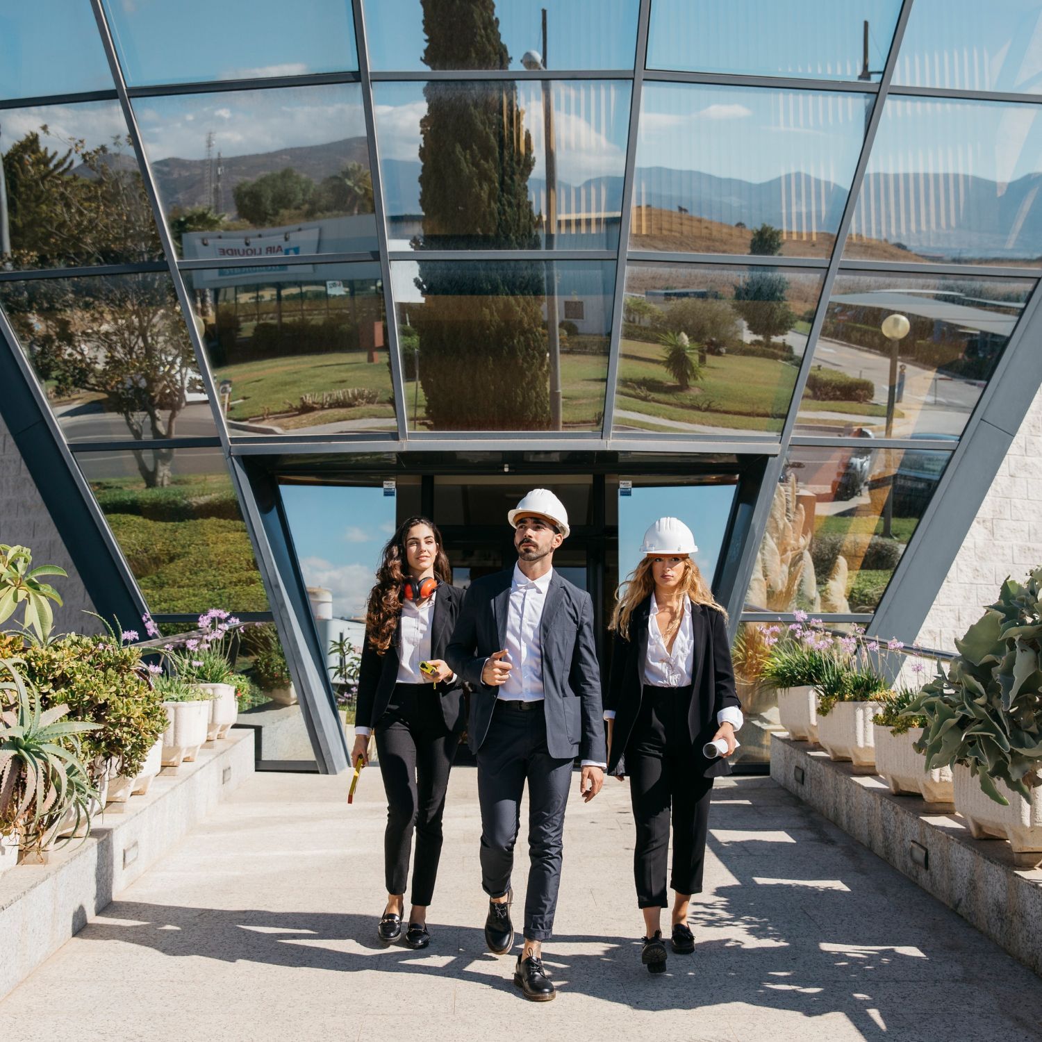 Three people wearing business suits and hard hats walk together from a modern glass-fronted building toward the camera.