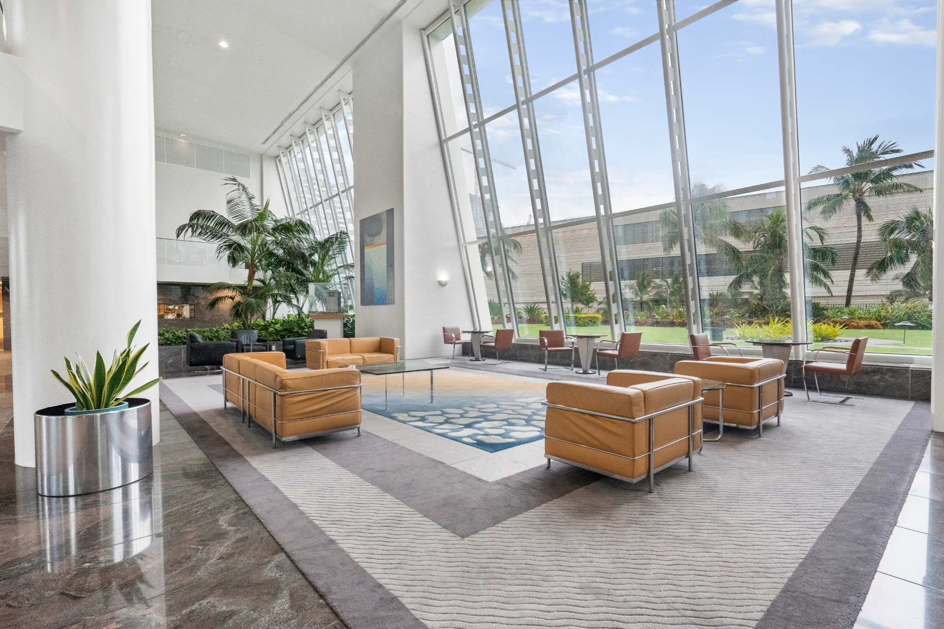 Spacious hotel lobby with tan leather chairs, large windows, a blue area rug, and indoor plants.